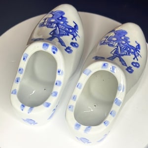 May include: Pair of white ceramic clogs with blue floral and windmill designs. The clogs have a rectangular opening and are decorated with blue squares around the edges. A classic Dutch souvenir.
