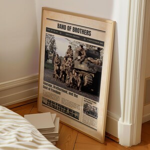 Pode incluir: Uma impress&atilde;o emoldurada com design de jornal, intitulada "BAND OF BROTHERS". A imagem apresenta uma fotografia a preto e branco de soldados e um tanque. O texto inclui "Courage, Brotherhood, and the Cost of Freedom."