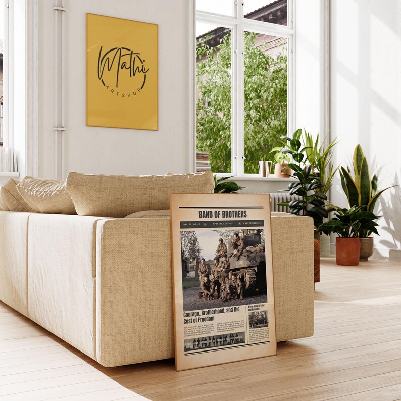 Pode incluir: Uma impress&atilde;o emoldurada de um artigo de jornal intitulado "Band of Brothers" com uma fotografia a preto e branco de soldados. A impress&atilde;o est&aacute; encostada a um sof&aacute; bege numa sala bem iluminada. Uma obra de arte emoldurada amarela est&aacute; pendurada na parede.