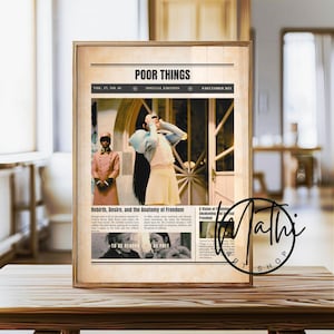 May include: A framed print featuring a newspaper-style design with the title "POOR THINGS." The image includes a central photo of a woman in a light-colored dress. Text includes "Rebirth, Desire, and the Anatomy of Freedom."