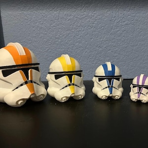 May include: A row of six white and black toy helmets with coloured stripes. The helmets feature orange, yellow, blue, purple, and red details. They are displayed on a black surface.