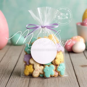 May include: A transparent cellophane bag tied with a lilac ribbon, filled with colourful, iced sugar biscuits. The biscuits are in various shapes and colours, including yellow, blue, and purple. The bag has a white circle for a logo and the text "1:1 Mockup".