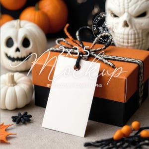 Puede incluir: Una maqueta con temática de Halloween con una caja de regalo naranja y negra con una etiqueta blanca en blanco. La caja está atada con hilo negro y naranja. En el fondo hay calabazas decorativas y una calavera. La etiqueta dice "2x3.5 Mockup".