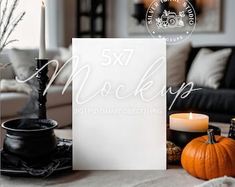 Op de afbeelding: Een 5x7 mockup met het woord "Mockups" in een scriptlettertype. De scène omvat een brandende kaars, een kleine pompoen en een zwarte ketel. De achtergrond toont een gezellige woonkamer.