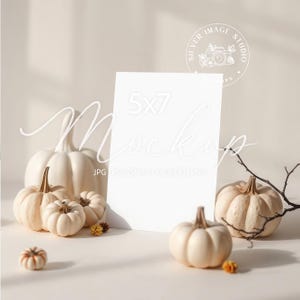 May include: A 5x7 mockup card stands among a collection of white and cream-colored pumpkins of various sizes. The pumpkins are arranged on a neutral surface, with a few small dried flowers and a twig adding to the autumn theme. The image has a soft, warm aesthetic.