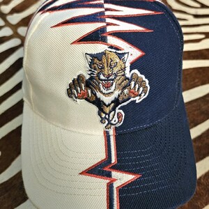 May include: A baseball cap featuring a split design with one side in cream and the other in navy blue. An embroidered panther graphic is centered on the front, accented by a jagged, lightning-like red and white line pattern running vertically.