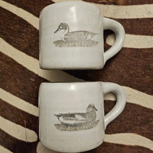 Peut inclure: Deux tasses en céramique blanc cassé avec une finition émaillée craquelée brillante. Chaque tasse présente une illustration monochrome détaillée d'un canard posé sur un sol texturé. Les tasses ont une anse épaisse et arrondie sur le côté.