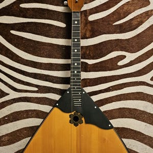 May include: A triangular balalaika string instrument featuring a light brown wooden body, a long neck with a dark fretboard, and a black pickguard surrounding a central circular sound hole with a decorative flower-shaped wooden inlay.
