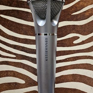May include: A silver Sennheiser XD48-U microphone with a mesh head. The microphone has a cylindrical body and a silver mesh head. The Sennheiser logo is printed vertically on the body. The background is a brown and white zebra print.