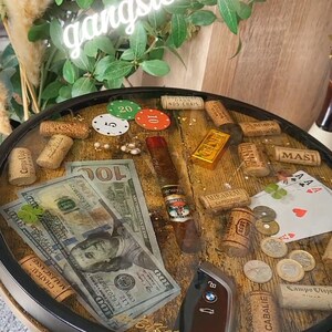 Op de afbeelding: Ronde tafel met een houten blad, versierd met pokerchips, bankbiljetten, een sigaar en wijnkurken. De tafel heeft een zwart metalen frame en poten. De tekst "gangster style" staat in een neonbord erboven.