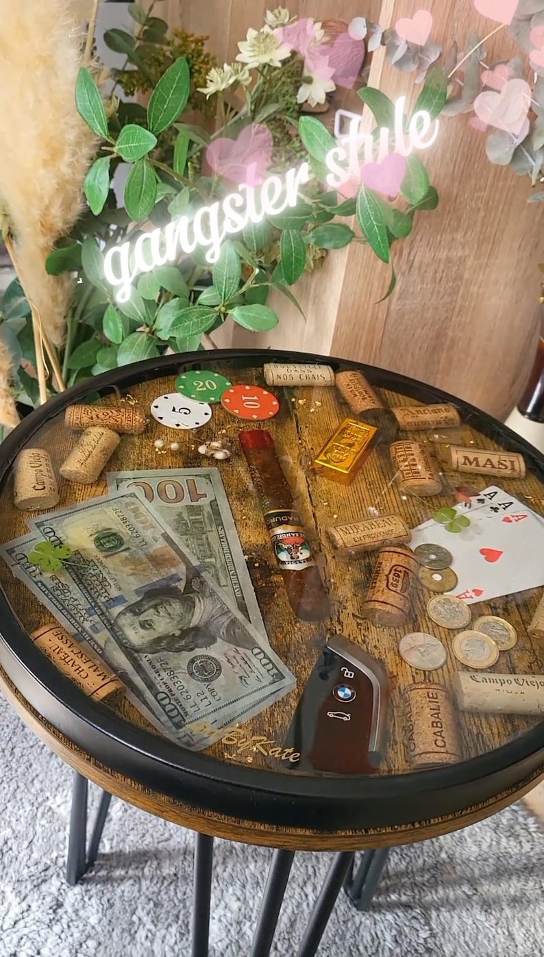 Op de afbeelding: Ronde tafel met een houten blad, versierd met pokerchips, bankbiljetten, een sigaar en wijnkurken. De tafel heeft een zwart metalen frame en poten. De tekst "gangster style" staat in een neonbord erboven.