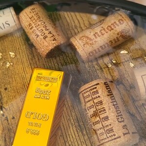 Op de afbeelding: Een goudstaaf met de tekst "GOLD" en "FINE 999.9" wordt weergegeven naast drie wijnkurken. De kurken hebben bedrukte tekst en de goudstaaf is gemarkeerd met "NET WT 200g". De achtergrond heeft goudvlokken.
