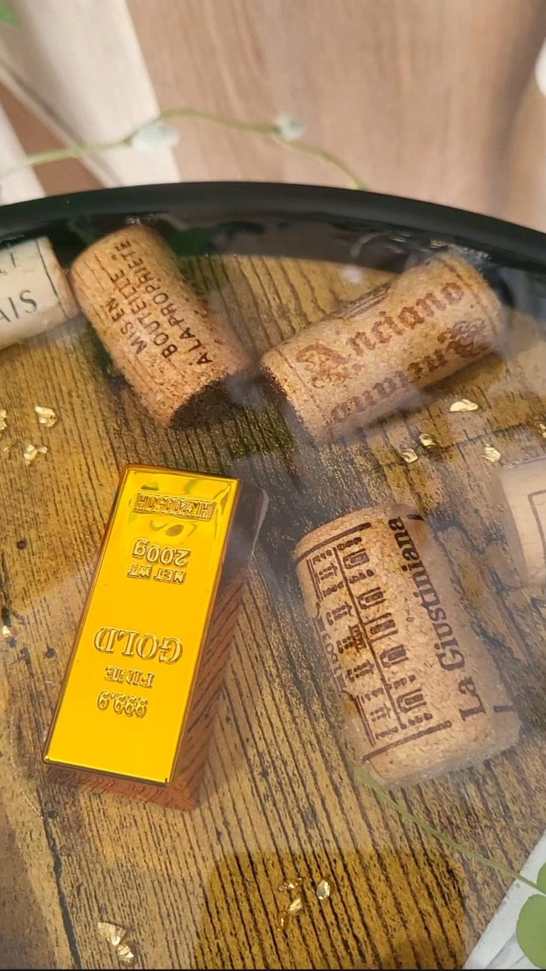 Op de afbeelding: Een goudstaaf met de tekst "GOLD" en "FINE 999.9" wordt weergegeven naast drie wijnkurken. De kurken hebben bedrukte tekst en de goudstaaf is gemarkeerd met "NET WT 200g". De achtergrond heeft goudvlokken.