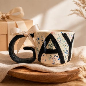 May include: A ceramic mug with the word "GAY" in bold black letters. The mug has a speckled design with blue, pink, and green accents. The handle is black and the mug is sitting on a wooden tray. A gift box with a bow is in the background.