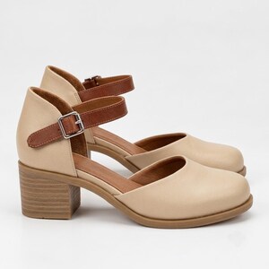 May include: A pair of beige shoes with a rounded toe, a block heel, and a brown ankle strap featuring a silver-toned metal buckle. The shoes have a smooth finish and a tan interior lining, showcasing a classic design with a sturdy heel construction.