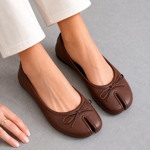 Könnte beinhalten: Ein Paar dunkelbraune Leder-Ballerinas mit geteilter Zehenpartie, abgerundeter Vorderkappe mit einem markanten vertikalen Schlitz und einer kleinen dekorativen Schleife auf dem Vorderblatt. Die Schuhe haben ein flaches Design mit glatter Oberfläche.