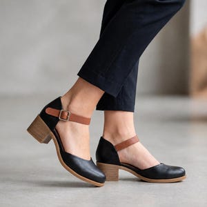 May include: Black leather shoes with a rounded toe, a tan ankle strap featuring a silver-toned metal buckle, and a light brown block heel. The shoes are paired with dark navy blue trousers that end above the ankle, showing the shoe design.