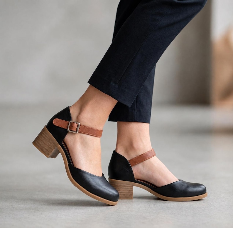 May include: Black leather shoes with a rounded toe, a tan ankle strap featuring a silver-toned metal buckle, and a light brown block heel. The shoes are paired with dark navy blue trousers that end above the ankle, showing the shoe design.