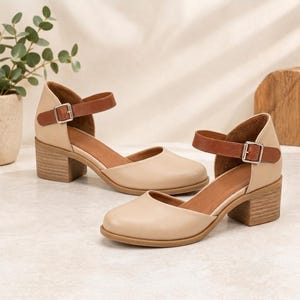 May include: A pair of beige shoes with a rounded toe, a low block heel, and a brown ankle strap featuring a silver-toned metal buckle. The shoes have a smooth finish and a cutout design on the sides, providing a classic look with a secure fit.