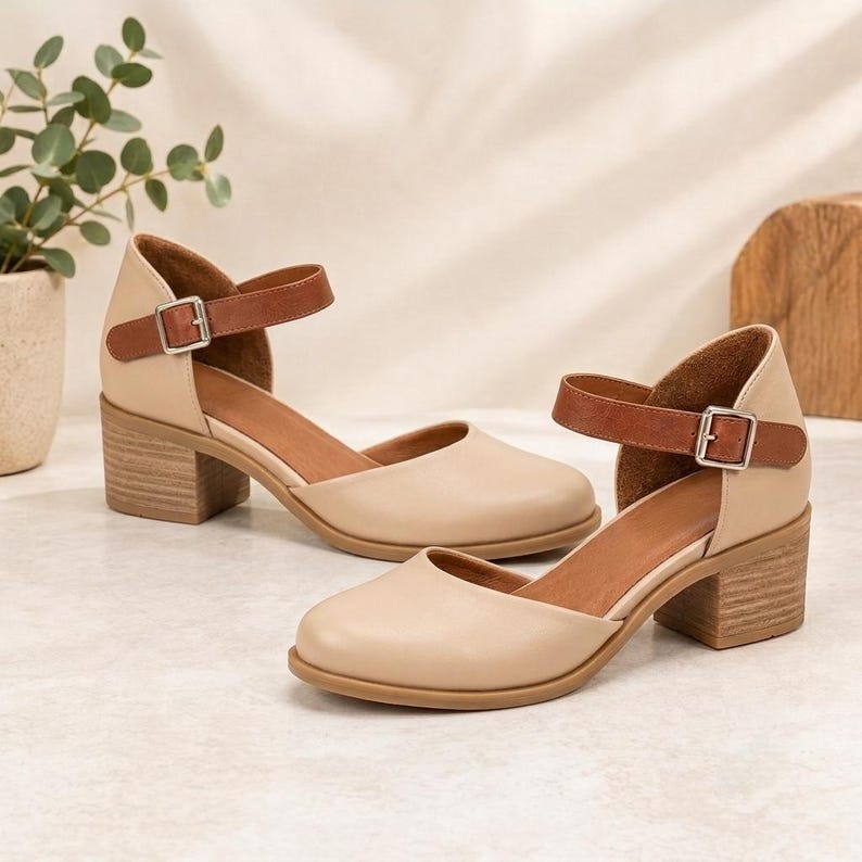 May include: A pair of beige shoes with a rounded toe, a low block heel, and a brown ankle strap featuring a silver-toned metal buckle. The shoes have a smooth finish and a cutout design on the sides, providing a classic look with a secure fit.