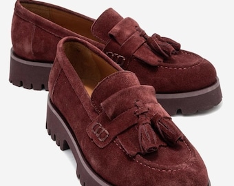 May include: A pair of burgundy suede loafers with a chunky sole. Each shoe features a decorative strap with a tassel and a stitched vamp. The shoes have a classic loafer design with a modern, elevated platform.
