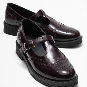 May include: A pair of burgundy Mary Jane style shoes with a T-strap and buckle closure. The shoes have a glossy finish, decorative perforations, and a thick black sole.