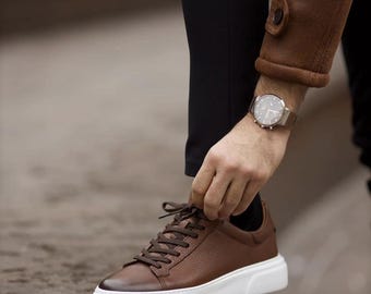 Puede incluir: Una zapatilla de cuero marrón con acabado texturizado, cordones marrón oscuro y una suela de goma blanca gruesa. El calzado presenta una punta redondeada con un sutil efecto degradado, combinado con pantalones negros y una manga de chaqueta.