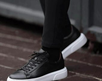 May include: Black leather sneakers with white soles and black laces. The shoes have a low-top design and are worn with black pants. The sneakers are a stylish and modern footwear choice.