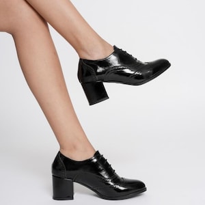 May include: Black patent leather oxford shoes with a block heel. The shoes have a classic wingtip design with decorative perforations and lace-up closure. The heels are approximately 3 inches high.