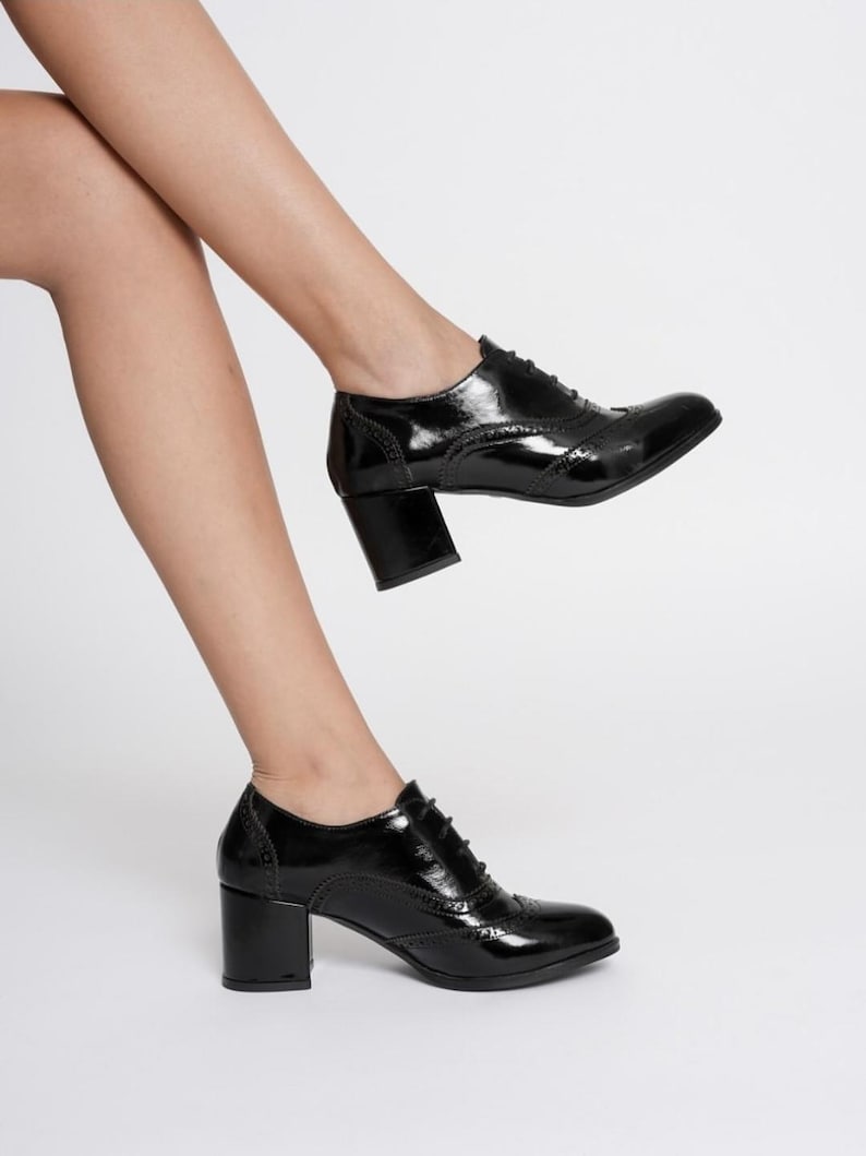 May include: Black patent leather oxford shoes with a block heel. The shoes have a classic wingtip design with decorative perforations and lace-up closure. The heels are approximately 3 inches high.