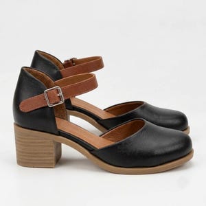 May include: A pair of black shoes with a rounded toe, featuring a tan ankle strap with a silver buckle and a block heel. The interior lining is tan, and the outer construction is black with a tan sole, providing a two-tone appearance.