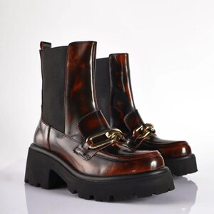 May include: A pair of brown and black ankle boots with a chunky black sole. The boots have a glossy finish and a gold-colored chain detail across the top. The boots have a pull-on design with black elastic side panels.