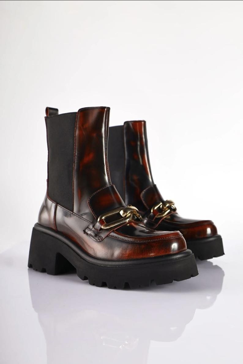 May include: A pair of brown and black ankle boots with a chunky black sole. The boots have a glossy finish and a gold-colored chain detail across the top. The boots have a pull-on design with black elastic side panels.
