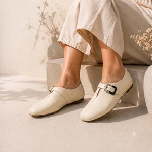 May include: A pair of cream-colored leather flats featuring a rounded toe, a central vertical seam, and a single strap across the instep secured with a rectangular buckle that has a dark, contrasting border and a metallic silver-toned center.
