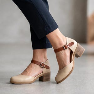 May include: A pair of beige leather shoes with a rounded toe, a brown ankle strap featuring a silver-toned metal buckle, and a stacked wooden block heel. The shoes have a cut-out design on the sides and are worn with dark navy blue trousers.