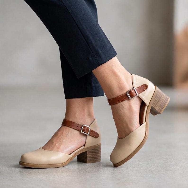 May include: A pair of beige leather shoes with a rounded toe, a brown ankle strap featuring a silver-toned metal buckle, and a stacked wooden block heel. The shoes have a cut-out design on the sides and are worn with dark navy blue trousers.