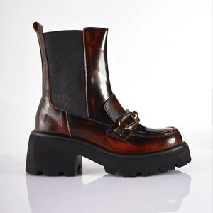 May include: Brown and black ankle boots with a chunky black sole. The boots have a glossy finish with a gold-colored chain detail on the front. The heel is approximately 3 inches high.