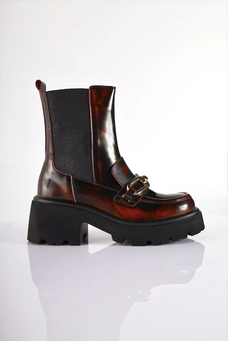 May include: Brown and black ankle boots with a chunky black sole. The boots have a glossy finish with a gold-colored chain detail on the front. The heel is approximately 3 inches high.