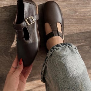 May include: A pair of dark brown T-strap shoes featuring a smooth finish, a rounded toe, and a gold-toned metal buckle closure on the side strap. The shoes have a low profile and a flat sole, showcasing a classic design with a secure ankle strap.