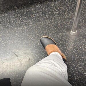 May include: A black shoe with a rounded toe and a tan ankle strap, worn with white pants. The shoe features a low heel and a visible buckle on the side. The overall design is simple and functional, showcasing a contrast between the dark upper and the light-colored trousers in a casual setting.