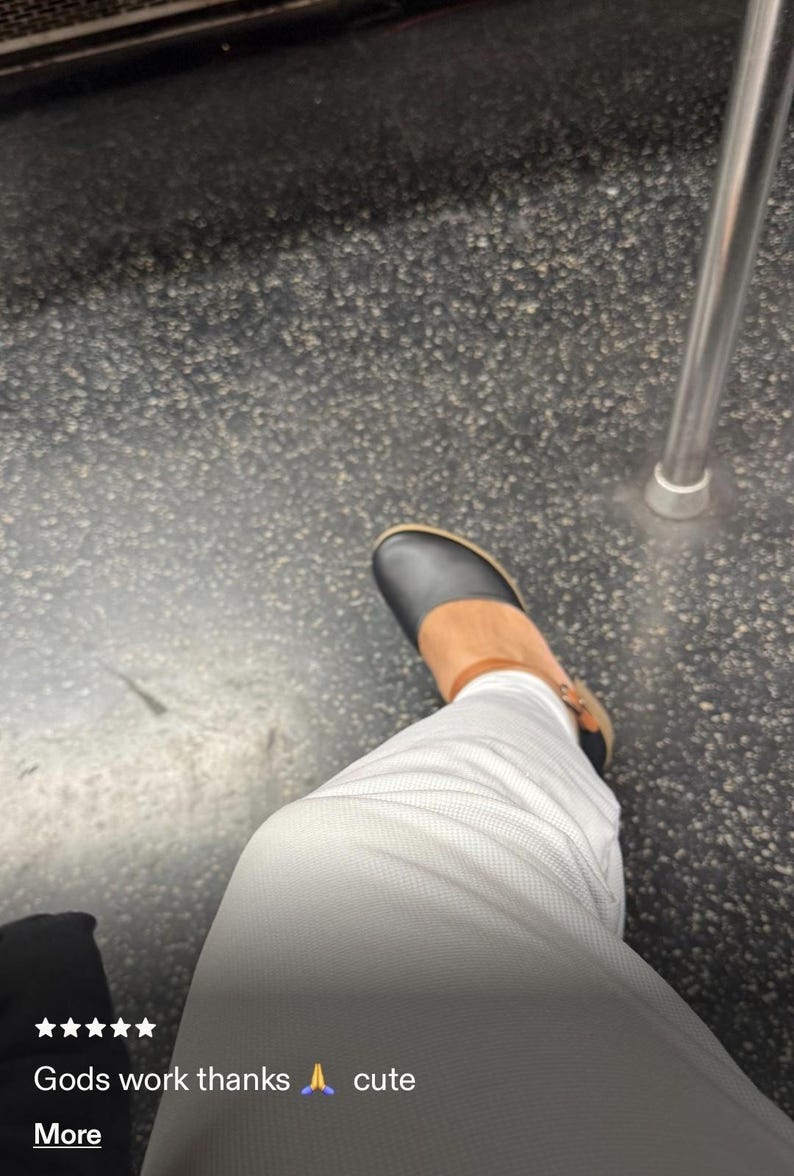 May include: A black shoe with a rounded toe and a tan ankle strap, worn with white pants. The shoe features a low heel and a visible buckle on the side. The overall design is simple and functional, showcasing a contrast between the dark upper and the light-colored trousers in a casual setting.