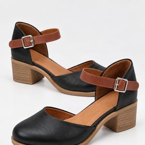 May include: A pair of black shoes with a rounded toe, featuring a tan ankle strap with a silver buckle and a block heel. The interior lining is tan, and the outsole is a light brown color. The design includes a cutout side detail.