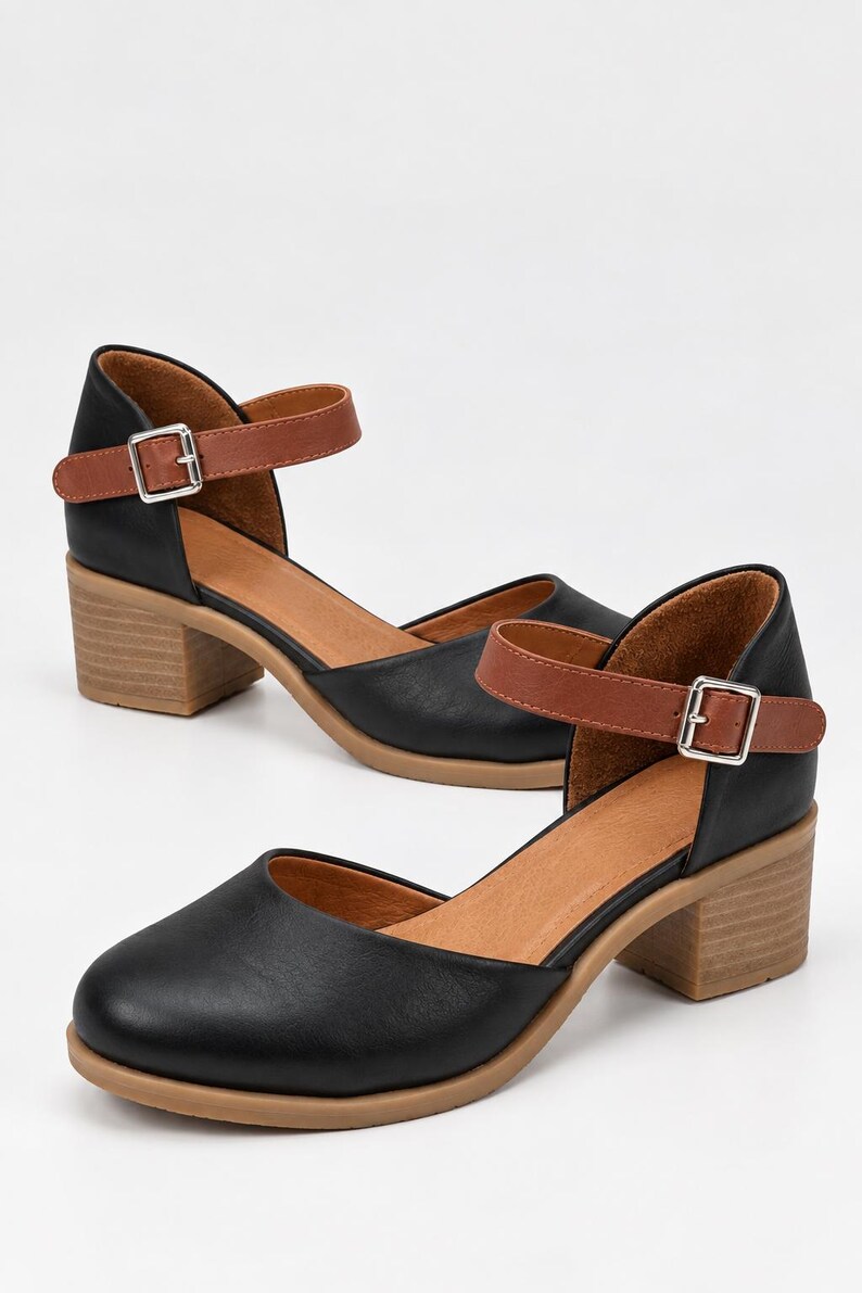 May include: A pair of black shoes with a rounded toe, featuring a tan ankle strap with a silver buckle and a block heel. The interior lining is tan, and the outsole is a light brown color. The design includes a cutout side detail.