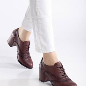 May include: Burgundy leather oxford-style shoes with a block heel. The shoes feature decorative perforations and black laces. The image shows the shoes being worn with white pants.