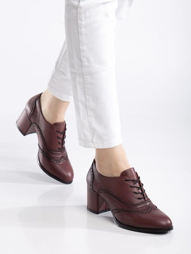 May include: Burgundy leather oxford-style shoes with a block heel. The shoes feature decorative perforations and black laces. The image shows the shoes being worn with white pants.