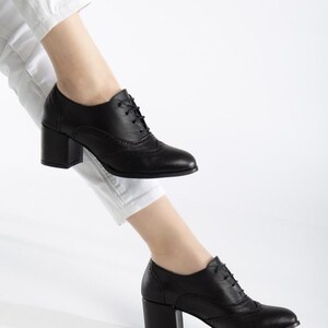 May include: Black leather oxford-style shoes with a block heel. The shoes feature a lace-up design and decorative perforations along the seams. The heel height is approximately 3 inches.
