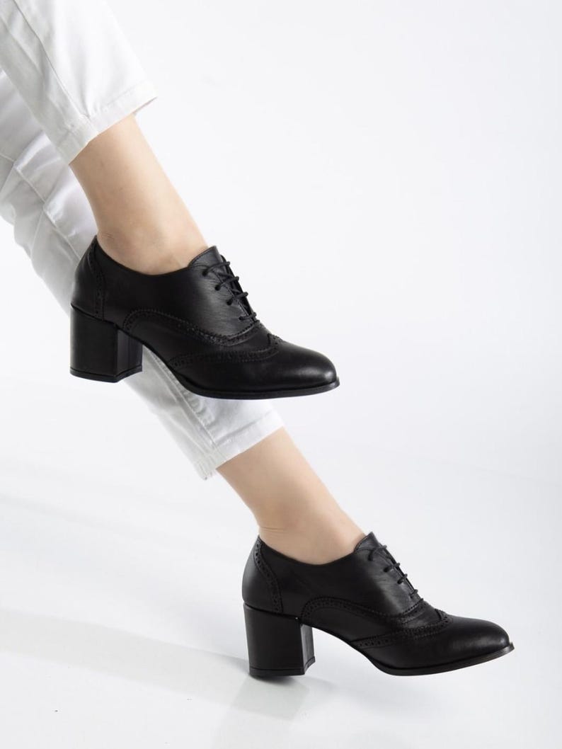 May include: Black leather oxford-style shoes with a block heel. The shoes feature a lace-up design and decorative perforations along the seams. The heel height is approximately 3 inches.