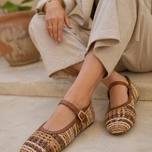 May include: Woven flat shoes with brown leather straps and gold buckles. The shoes have a tan and brown woven pattern. The shoes are a Mary Jane style. The shoes are on a light-colored surface.