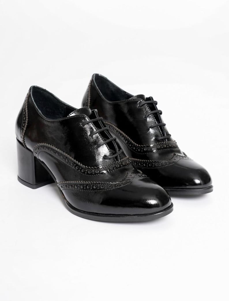 May include: A pair of black patent leather Oxford shoes with a block heel. The shoes have a classic lace-up design and decorative brogue detailing along the edges. The heels measure approximately 2.5 inches.