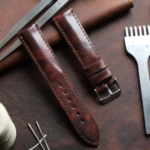 Könnte beinhalten: Zwei braune Lederarmbänder mit strukturierter Oberfläche. Die Armbänder haben ein klassisches Design mit Nähten an den Kanten und einer silberfarbenen Schnalle. Das Bild zeigt auch Werkzeuge für Lederarbeiten.